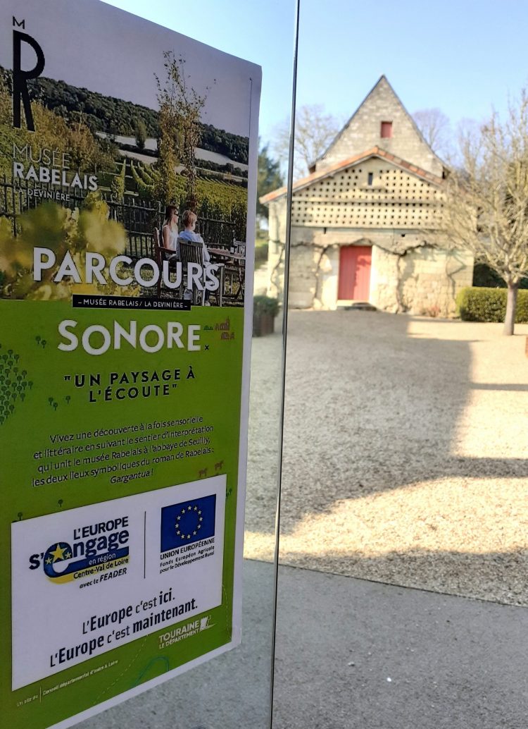 En premier plan, l'affiche du parcours sonore du musée Rabelais attestant de son cofinancement par LEADER. En arrière plan, la maison de la Devinière avec une porte rouge et un petit arbre à droite